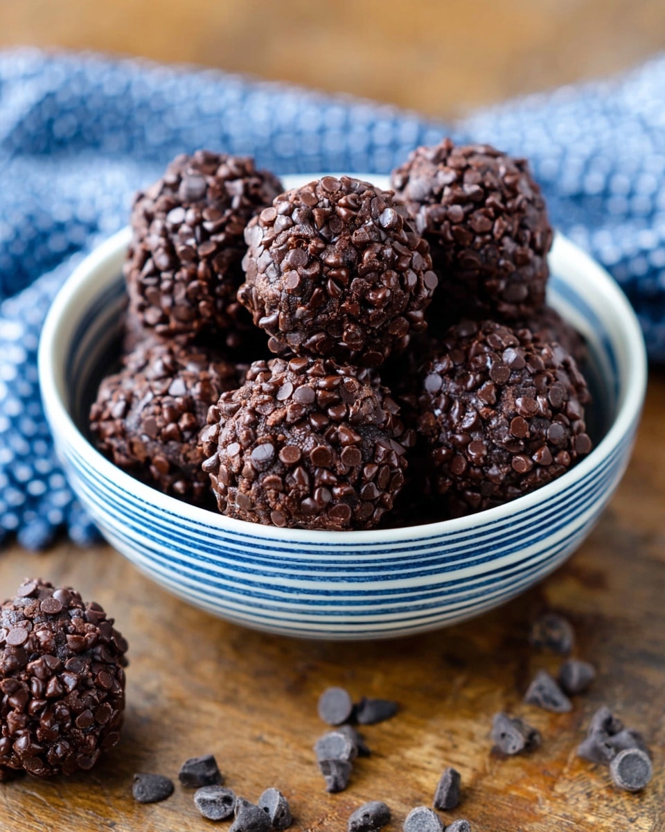 A white bowl filled with about ten round chocolate cookie balls, each covered densely in small, dark chocolate chips, giving them a rough, textured surface. The cookies have a rich, deep brown color peeking through the scattered chips, and they sit close together in the bowl. The bowl has thin blue stripes running vertically on the inside. Around the bowl, a few chocolate chips are scattered on a white marbled surface. A blue and white checkered cloth is softly blurred in the background. photo taken with an iphone --ar 4:5 --v 7