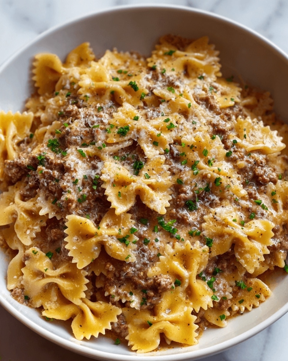 A white bowl filled with about two layers of farfalle pasta mixed with ground beef. The pasta is creamy light yellow with a smooth texture and peppered with small bits of dark brown cooked beef. The dish is topped with a sprinkle of finely chopped green herbs and black pepper, creating small specks of color across the surface. The creamy sauce coats the pasta evenly, shining slightly in the light. The bowl sits on a white marbled surface. photo taken with an iphone --ar 4:5 --v 7