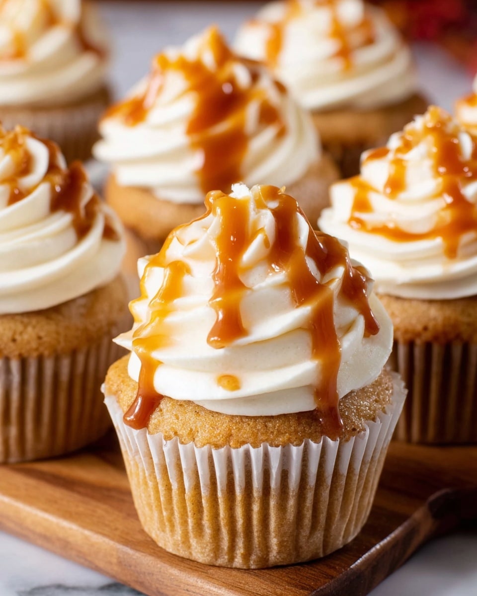 The image shows several cupcakes with three main layers: the base is a golden brown cupcake with ribbed white paper lining, held by a woman's hand. On top of the cupcake is a thick swirl of smooth, creamy white frosting with soft peaks. Drizzled over the frosting is a shiny caramel sauce with rich amber tones, flowing in thin, uneven lines. The cupcakes are arranged close together on a wooden surface with a blurred white marbled background. Photo taken with an iphone --ar 4:5 --v 7