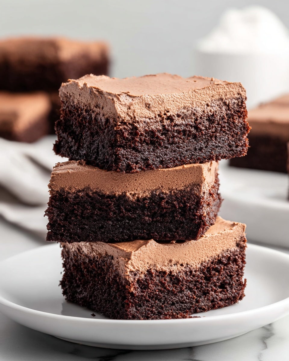 A stack of three chocolate brownies is shown on a white plate placed on a white marbled surface. Each brownie has two layers: a thick, dark brown, moist and dense bottom brownie layer, and a lighter brown, fluffy, smooth chocolate mousse layer spread evenly on top. The edges of the mousse layer have a slightly textured, whipped look. In the background, more brownies on a white plate and a white object are slightly blurred. Photo taken with an iphone --ar 4:5 --v 7