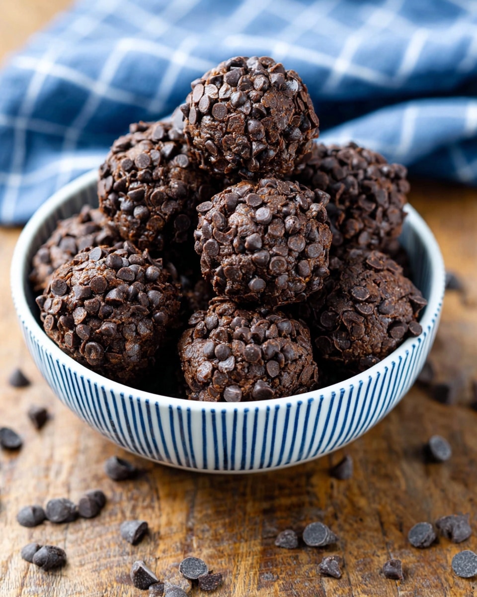 The image shows a white bowl with blue stripes filled with round chocolate balls covered in small dark chocolate chips. Each ball has a rough, textured surface fully coated with dense, shiny chocolate chips, making the balls look rich and moist. The bowl sits on a wooden surface with some scattered chocolate chips around it, and a blurred blue and white checkered cloth is in the background. photo taken with an iphone --ar 4:5 --v 7