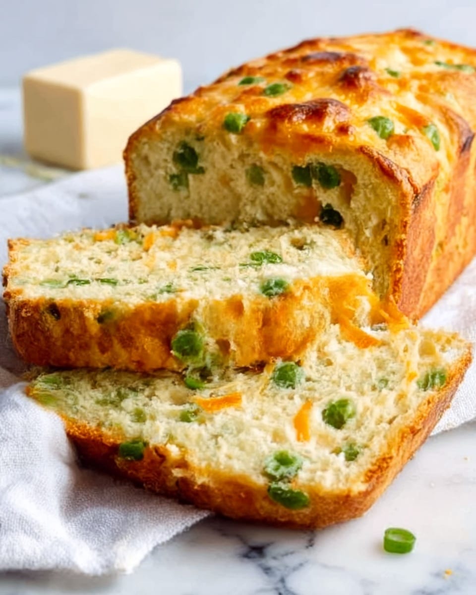 A loaf of savory bread is shown on a white marbled surface, with one thick slice cut and lying next to it. The bread has a golden-brown crust with a slightly rough, baked texture. Inside, the bread is light yellow with green pea pieces evenly spread throughout. The slice reveals a soft, airy crumb with occasional little holes, and a layer of melted orange cheese visible near the middle. In the background, there is a blurred white bowl with cubes of butter. Photo taken with an iphone --ar 4:5 --v 7