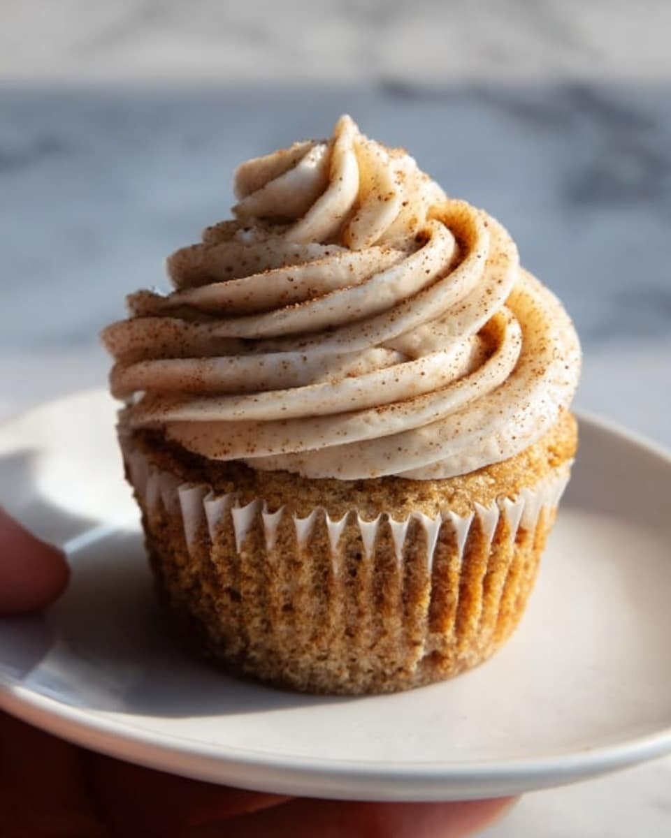 A single cupcake sits on a white plate, showing a soft golden-brown cake base with a light crumb texture. On top, there is a thick swirl of creamy, pale beige frosting with small specks sprinkled over it, possibly cinnamon or nutmeg. The frosting is smoothly piped in layers forming a tall peak with ridges that spiral upward. A woman's hand is about to touch the side of the cupcake. The background features a white marbled surface with a soft natural light illuminating the cupcake. Photo taken with an iphone --ar 4:5 --v 7