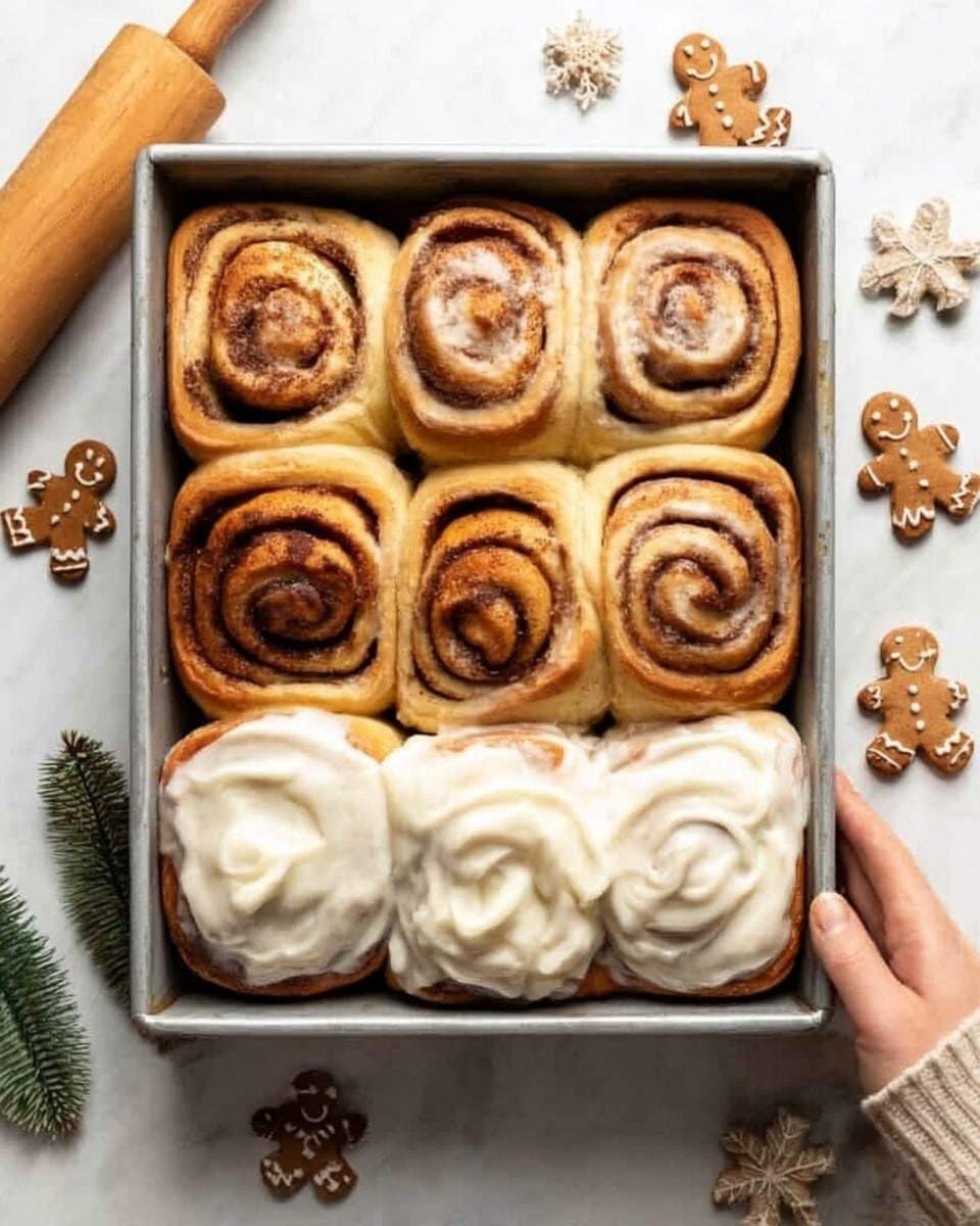 A metal rectangular baking tray filled with twelve cinnamon rolls arranged in three rows of four. The top two rows have golden-brown rolls with swirled layers of cinnamon and sugar, their texture soft and slightly shiny from baked glaze. The bottom row contains four cinnamon rolls covered in creamy white icing, thick and smooth, partially melting into the layers of dough beneath. The tray sits on a white marbled surface with a wooden rolling pin on the left and small gingerbread cookies scattered nearby. A woman's hand is gently placed near the top right corner of the tray. photo taken with an iphone --ar 4:5 --v 7