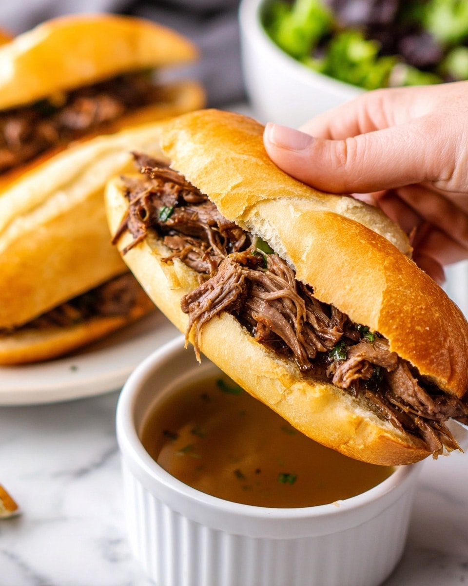 A close-up view of a sandwich held by a woman's hand, showing a soft, golden brown toasted roll split open and filled with a generous layer of shredded dark brown meat, which looks juicy and tender. The sandwich is dipped into a white small bowl filled with a light brown broth, and another similar sandwich is visible in the background resting on the white marbled surface. In the blurred background, there is a white bowl containing green and dark leafy salad. photo taken with an iphone --ar 4:5 --v 7