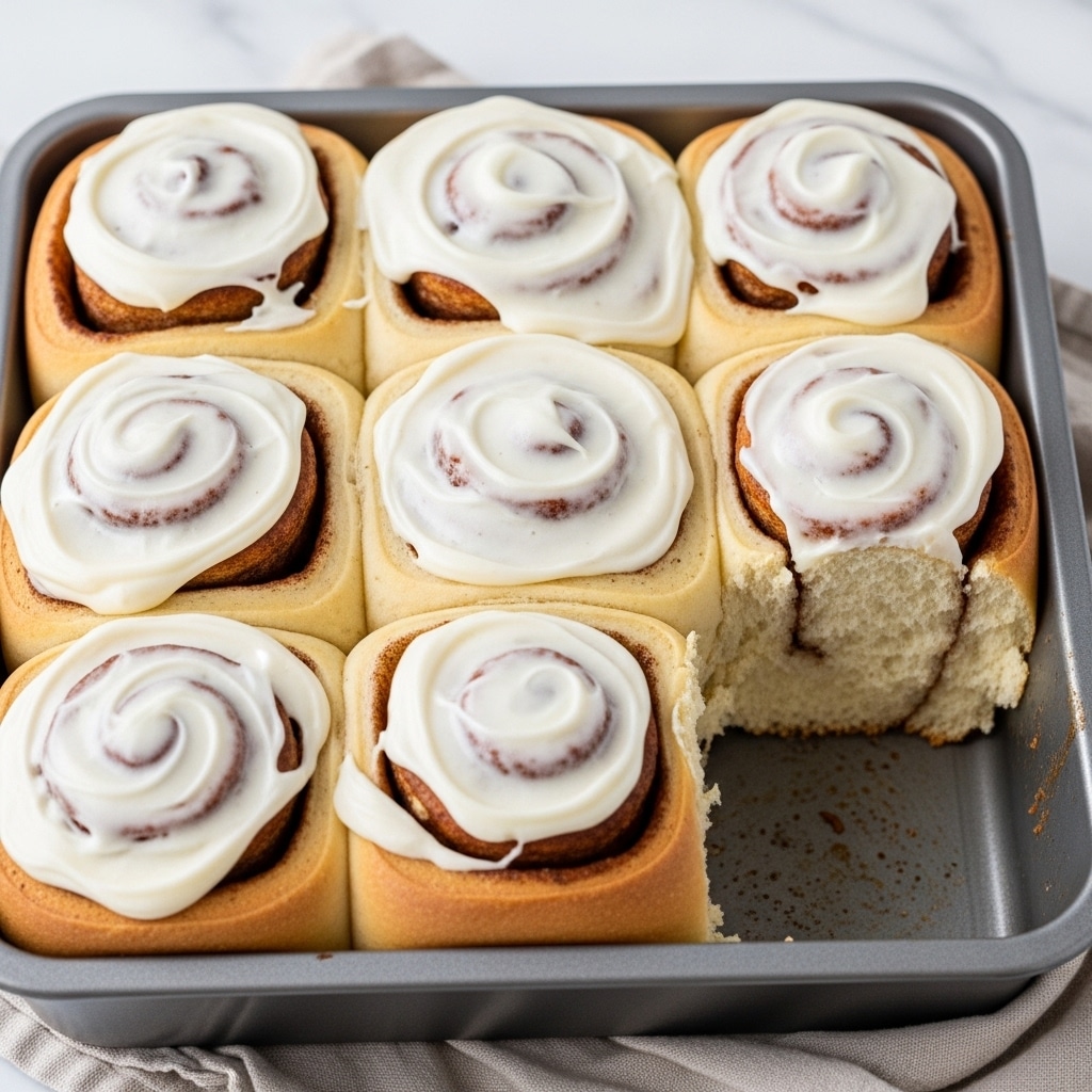 A metal baking pan filled with eight soft, thick cinnamon rolls, each with a visible spiral layer of golden-brown dough and cinnamon filling. The rolls are topped with a creamy, white frosting that is swirled unevenly, dripping slightly on the sides. The texture of the dough looks fluffy and light, with a slightly toasted golden edge on the bottom. One cinnamon roll is pulled away from the group, showing the soft and airy inside. The pan sits on a white marbled surface with a cloth partially underneath. photo taken with an iphone --ar 4:5 --v 7