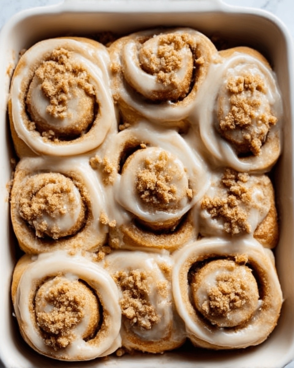 A white rectangular baking dish filled with eight cinnamon rolls arranged closely in two rows, each roll showing a soft, golden-brown spiral shape covered with a thick layer of beige icing that looks smooth and glossy. The tops of the rolls have small clusters of crumbly light brown streusel sprinkled unevenly. The dish is on a white marbled surface, and a woman's hand is visible holding the dish from the side. photo taken with an iphone --ar 4:5 --v 7