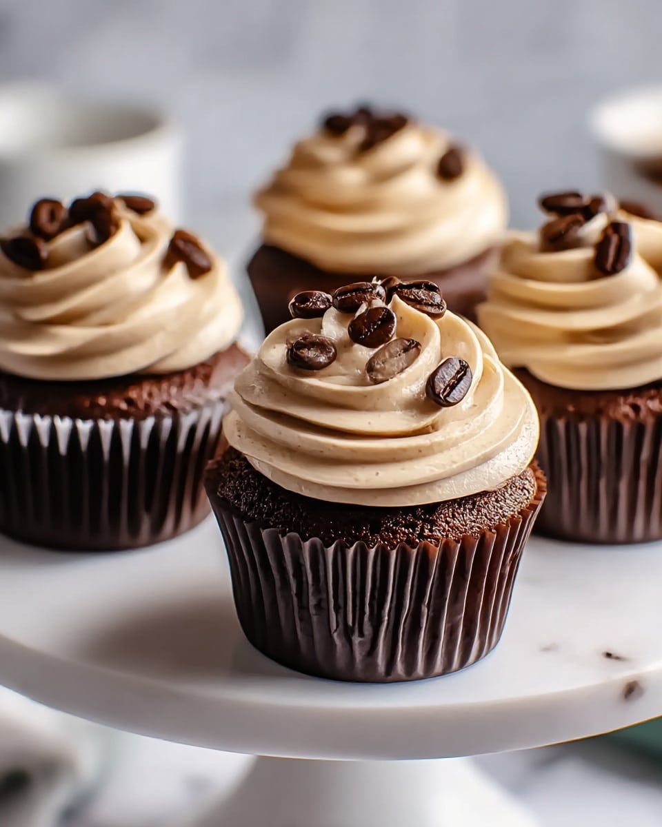 A close-up of four chocolate cupcakes on a white cake stand. Each cupcake has a dark brown base with a rich texture, wrapped in a dark brown paper liner, topped with a thick swirl of creamy light brown frosting that looks smooth and fluffy. The frosting is decorated with shiny dark brown coffee beans scattered on top. The background and surface are a white marbled texture, softly blurred to keep focus on the cupcakes. Photo taken with an iphone --ar 4:5 --v 7