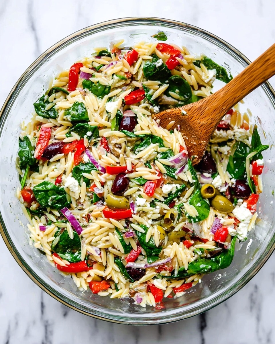 A clear glass bowl filled with a colorful salad sits on a white marbled surface. The salad has four main layers: a base of pale yellow orzo pasta shaped like small grains, scattered fresh dark green spinach leaves, chopped red bell peppers and red onion pieces adding splashes of bright red and purple, and halved green and black olives dotted throughout. Some white crumbled feta cheese pieces are mixed in, giving a creamy contrast. A wooden spoon stands inside the bowl on the right side, partially stirring the ingredients. Photo taken with an iphone --ar 4:5 --v 7