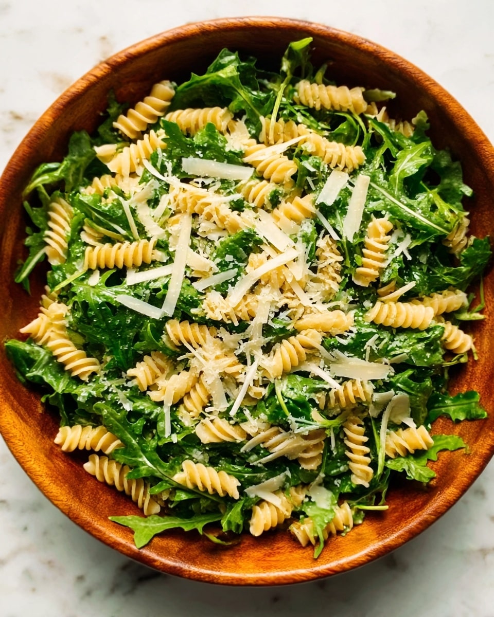 A white bowl filled with a fresh salad showing three main layers: the bottom layer is dark green leafy arugula, the middle layer is light green chopped herbs scattered evenly, and the top layer is spiral-shaped pale yellow pasta pieces spread across, mixed with thin, light beige cheese slices. The textures range from soft leaves to chewy pasta and firm cheese. Everything sits on a white marbled surface. Photo taken with an iphone --ar 4:5 --v 7