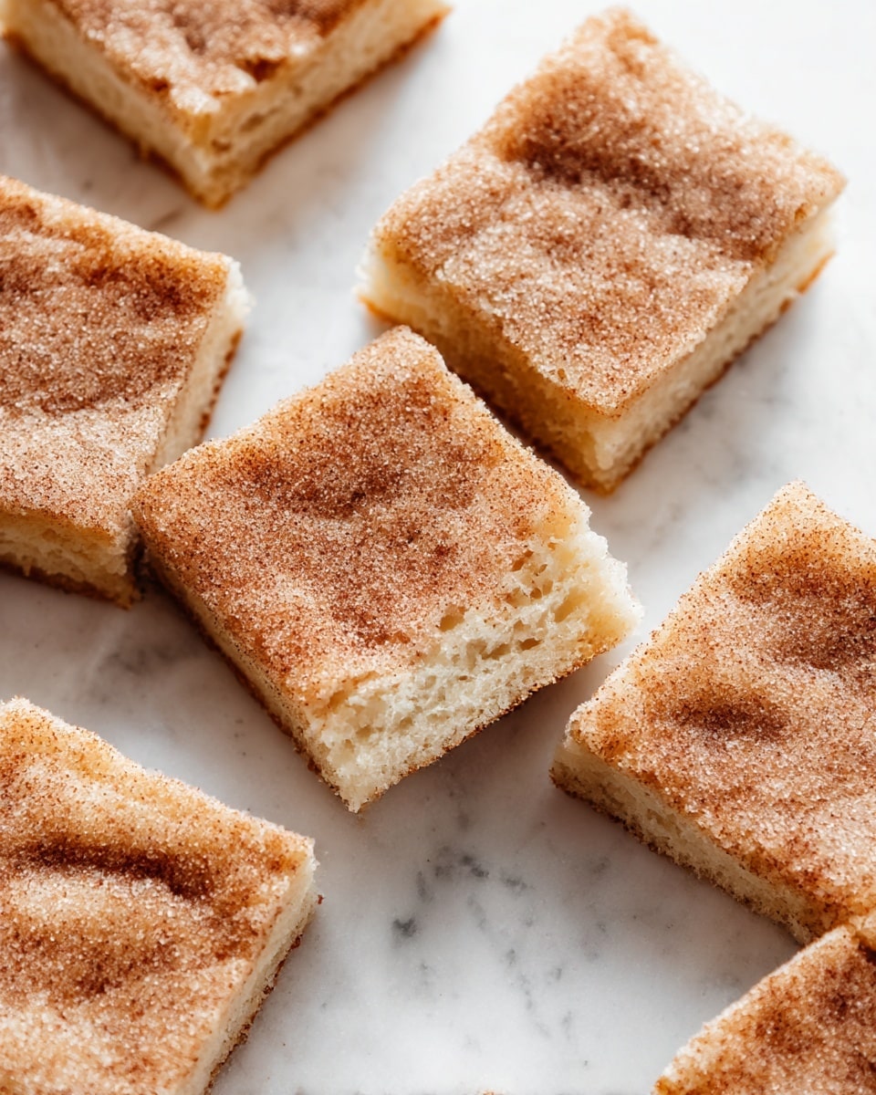 The image shows multiple squares of soft, light beige sponge cake with a generous topping of cinnamon and sugar, giving a textured, slightly grainy top layer that is darker brown with a sparkling sugar coating. Each square piece is stacked or laid closely together on a crinkled white parchment paper, placed on a white marbled surface. The cake layers are thick and fluffy, with the cinnamon sugar evenly spreading across the surface, creating a rich contrast between the light cake base and the cinnamon topping. The overall appearance is warm and inviting, with a simple homemade look. photo taken with an iphone --ar 4:5 --v 7