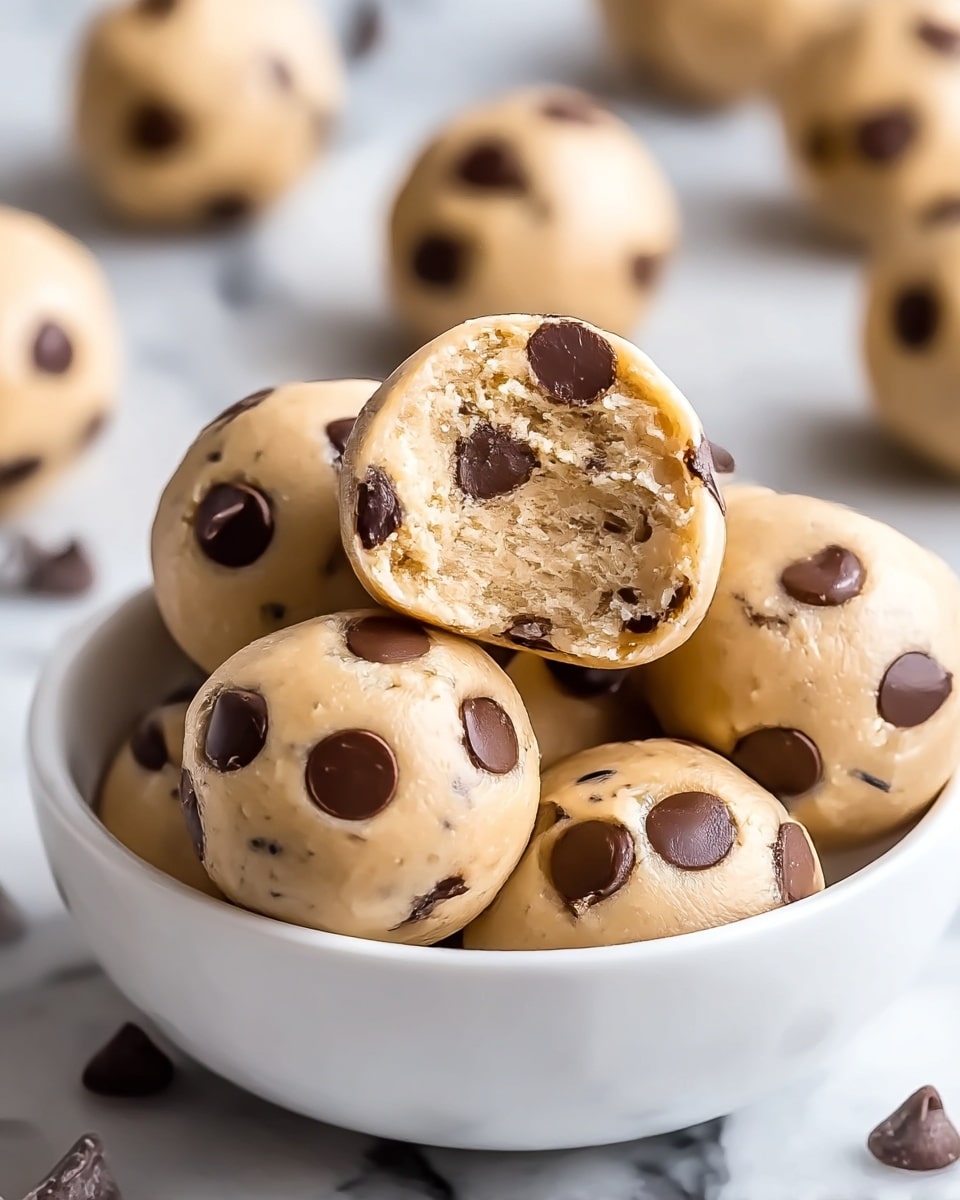 The image shows a white bowl filled with round cookie dough balls studded with large, dark brown chocolate chips. One dough ball is broken in half on top, revealing a soft, beige dough interior with embedded chocolate chips. The dough balls have a smooth texture with small black specks, possibly vanilla bean, scattered throughout. The bowl sits on a white marbled surface, with more dough balls and chocolate chips blurred in the background. Photo taken with an iphone --ar 4:5 --v 7