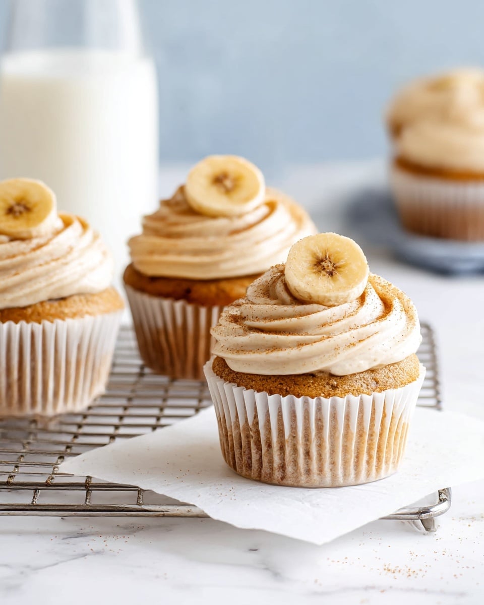 Four light brown cupcakes sit on a silver cooling rack over white paper on a white marbled surface. Each cupcake has a soft, swirled layer of creamy beige frosting on top, sprinkled lightly with cinnamon, and a dried banana chip is placed upright at the center of each frosting swirl. The cupcakes are wrapped in white paper liners. In the blurry background, there is a clear glass filled with milk and another cupcake. photo taken with an iphone --ar 4:5 --v 7