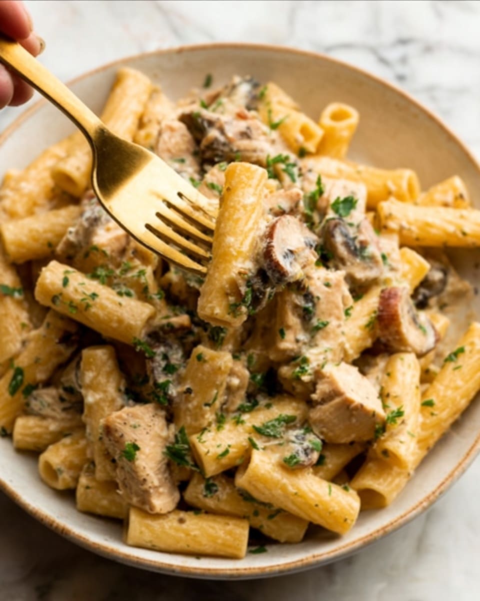 A white bowl filled with rigatoni pasta covered in creamy mushroom sauce, showing small pieces of browned chicken and sliced mushrooms mixed throughout. The creamy sauce coats the pasta giving it a light beige color, with green parsley sprinkled on top for a fresh look. A golden fork held by a woman's hand is piercing into the pasta, lifting some rigatoni. The bowl is set on a white marbled surface. photo taken with an iphone --ar 4:5 --v 7